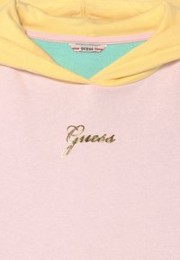 Pastel lyserød hoodie med gul hættefor, teal indvendig krave og gylden kursiv "Guess" logo på brystet.