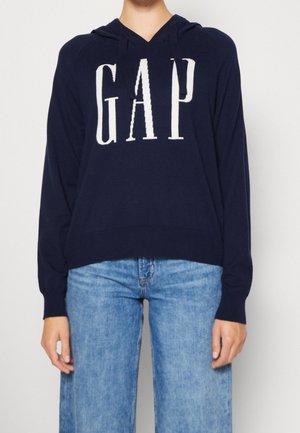 Personne portant un sweat à capuche bleu marine avec le texte « GAP » en blanc et un jean bleu clair, debout devant un fond clair uni.