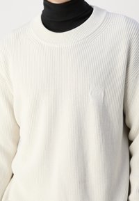 Bílý žebrovaný pletený svetr s kulatým výstřihem a malou nášivkou s logem na hrudi. Dlouhé rukávy a strukturovaná tkanina.
