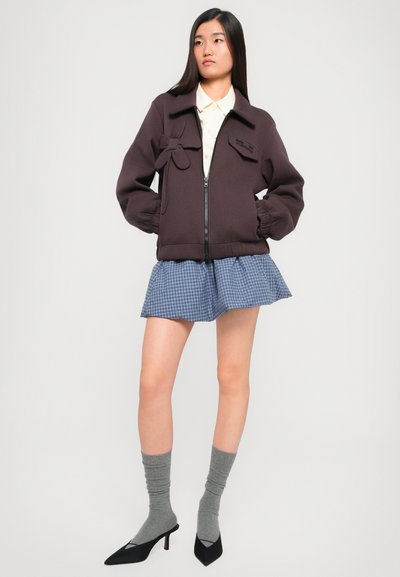 Chaqueta marrón con cremallera y puños elásticos, camisa de cuello debajo, falda a cuadros azul, medias gris hasta la rodilla y tacones puntiagudos negros.