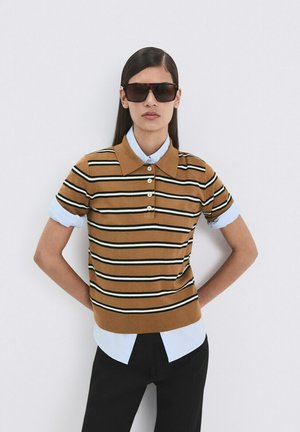 Kvinde med langt mørkt hår iført en oversize brunstribet polotrøje over en lyseblå skjorte med knapper, sorte bukser og store mørke solbriller, hænderne bag ryggen.