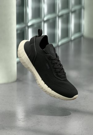 Zapatilla negra de malla con suela cremosa gruesa y con dibujo, flotando sobre un suelo gris en una habitación con ventanas de bloques de vidrio.