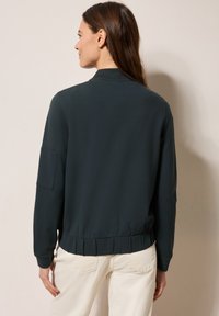 Giacca bomber verde scuro con orlo e polsini a coste, dotata di una piega sul retro e di una tasca laterale. Tessuto liscio con una vestibilità casual.