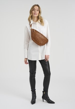 Naine, kes kannab valget särki, musti nahkseid pükse, musti pahkluu saapaid ja pruunist punutud risti keha kotti, helehallil ühtlasel taustal.