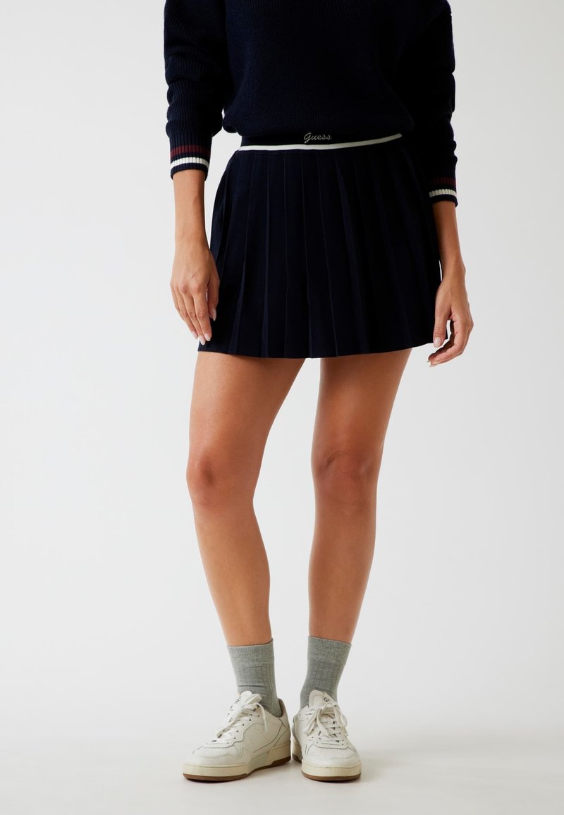 Marineblauer plissierter Rock mit einem weißen Bunddetail. Trägt graue Socken und weiße Sneaker. Glattes Material und klare Falten sichtbar.