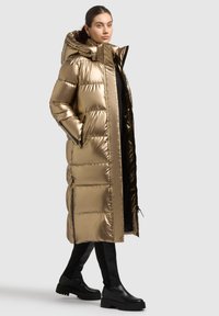 Pūkinis, metalinio aukso spalvos ilgas paltas su gaubtu, užtrauktuku ir šoninėmis kišenėmis. Turi siuvinėtą dizainą ir siekia žemiau kelio.