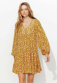 Robe florale jaune moutarde avec de longues manches bouffantes, décolleté en V et détails brodés. Présente une coupe ample et une jupe à volants avec un motif ludique.