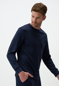 Maglietta a manica lunga blu navy in tessuto morbido, con scollatura rotonda e vestibilità slim. Il modello si fotografa con una mano in tasca, polsini testurizzati.