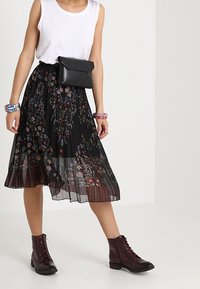 Jupe plissée noire à motif floral, associée à un débardeur blanc et un sac bandoulière noir. Des bottines en cuir bordeaux à laçage complètent le look.