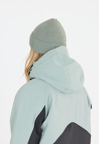 Vihreä vedenpitävä takki hupulla, jossa on mustia yksityiskohtia ja teksturoitu harmaa pipo. Takakuvasta näkymä designista ja materiaalista.