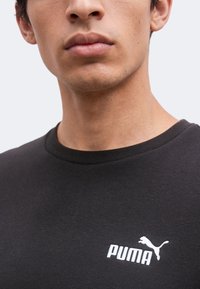 Jeune homme portant un t-shirt Puma noir avec logo blanc, visible du menton au haut de la poitrine sur un fond clair et uni.