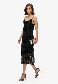 Robe midi en dentelle noire à fleurs avec bretelles spaghetti ajustables, ourlet inférieur transparent et doublure en satin lisse. Associée à une pochette transparente.
