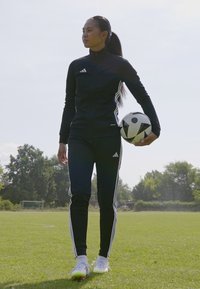 Veste et pantalon de sport noirs avec des rayures blanches, tenant un ballon de football noir et blanc, debout sur un terrain vert.