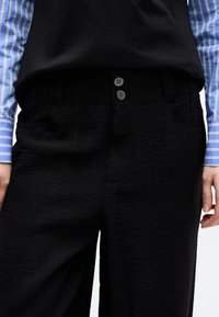 Personne portant un pantalon noir texturé à deux boutons et une chemise à manches longues rayée bleu et blanc, debout les mains détendues le long du corps.