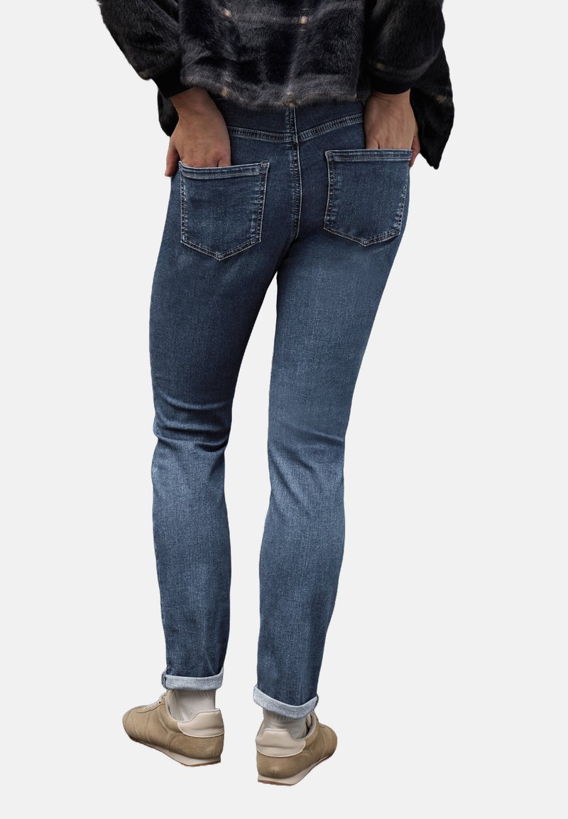 Pantalones de mezclilla azul oscuro con forma ajustada, que cuentan con dos bolsillos traseros y puños enrollados. Llevados con zapatillas de color claro.