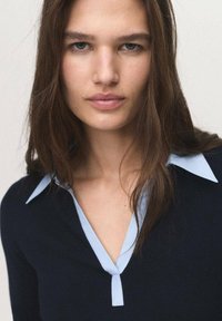 Young woman with long brown hair wearing a black sweater with a light blue collar, looking directly at the camera with a neutral expression.
