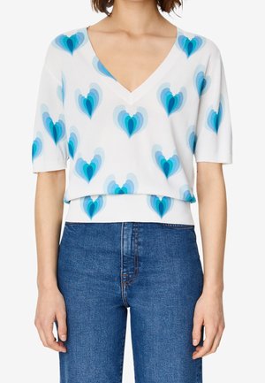 Femme portant un top blanc à col en V et manches courtes avec des motifs de cœurs bleus superposés, associé à un jean bleu taille haute.