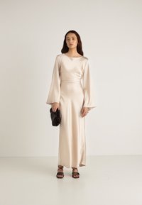 Satino smėlio spalvos ilga suknelė su varpo formos rankovėmis ir aptemptu liemeniu, derinama su juoda delninuke ir juodomis basutėmis su dirželiais. Glotni tekstūra.