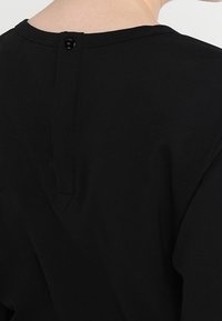 Černý bavlněný top s kulatým výstřihem a zapínáním na knoflík vzadu. Látka vypadá hladce s minimální texturou. Jednoduchý design.