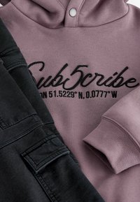 Sweat à capuche en doux lavande avec texte brodé noir, associé à un pantalon noir avec poches plaquées et tissu texturé.