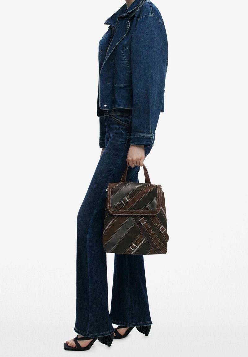 Personne portant une veste en jean bleu foncé et un jean, tenant un sac à main en cuir marron rayé avec des boucles, debout sur des sandales noires à talons.