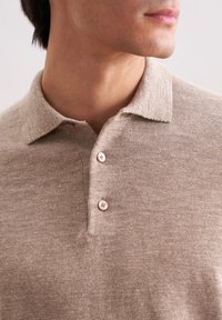 Mann trägt ein hellbraunes gestricktes Polohemd mit zwei befestigten Knöpfen, von den Schultern bis zum Mund vor einem schlichten Hintergrund gezeigt.