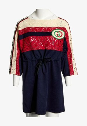 Robe avec jupe bleu marine, haut en dentelle rouge et crème, manches trois-quarts, ceinture noire à la taille et logo circulaire vert et rouge sur la poitrine.