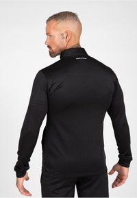 Zwarte sportieve shirt met lange mouwen, hoge kraag en een aansluitend ontwerp, gemaakt van rekbaar materiaal. Heeft de merknaam op de achterkant.