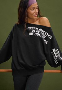 Felpa nera oversize con grafiche testuali bianche sulle maniche, caratterizzata da un colletto a costine e una vestibilità rilassata. Indossata con leggings scuri.