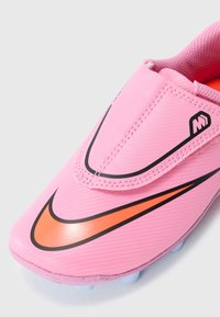 Ružová futbalová kopačka s oranžovým a čiernym Nike Swoosh, so zapínaním na suchý zips a hrotmi na bielej pozadí.