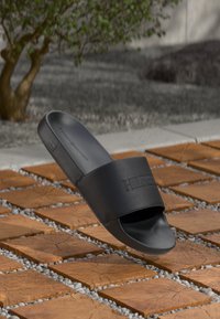 Single black slide sandal on wooden tile and pebble outdoor flooring with tree and concrete wall in the background.