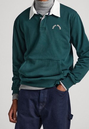 Mężczyzna ubrany w ciemnozieloną koszulkę polo z długim rękawem, białym kołnierzykiem i logo „Pepe Jeans”, szary golf pod spodem oraz ciemnoniebieskie spodnie, z ręką w kieszeni.