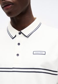 Άνδρας που φοράει λευκό πόλο με διπλές μπλε ρίγες στο γιακά και στο στήθος, κλειστό μπροστά με κουμπιά, με το λογότυπο Calvin Klein στο αριστερό στήθος.
