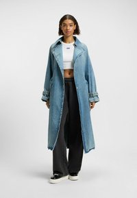 Manteau en denim, bleu clair, coupe ample, revers surdimensionné, boutons sur le devant, détail à l'ourlet brut, associé à un pantalon large noir et un haut blanc.