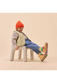 Ein Kind sitzt auf einem gesprenkelten Plastikhocker, trägt eine beige gesteppte Weste, eine orange Strickmütze und bunte, gemusterte Schuhe mit Klettverschlüssen.