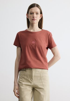 Junge Frau mit glatten braunen Haaren, die ein rostfarbenes T-Shirt mit Sonnenmotiv und eine beige Hose mit hohem Bund trägt, vor weißem Hintergrund stehend.