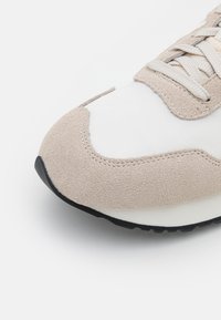 Lättviktig sneaker med ovandel i mocka och tyg i beige och vitt, som har en mjuk textur, snörning och en svart gummisul.