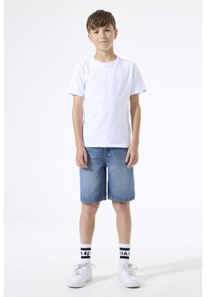 Jongen staand met ontspannen handen, draagt een wit T-shirt, spijkershorts, witte sneakers en witte sokken met zwarte tekst tegen een effen achtergrond.