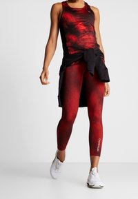 Czerwono-czarny sportowy top na ramiączkach z nakrapianym wzorem, zestawiony z czerwonymi legginsami i czarną bluzą przewiązaną w talii. Białe sneakersy.
