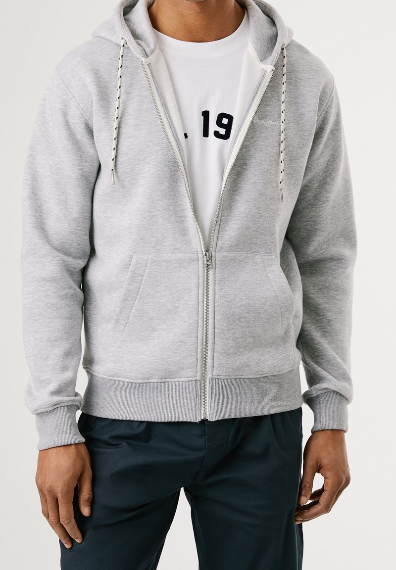 Homme portant un sweat à capuche gris clair zippé sur un t-shirt blanc avec un motif noir, assorti à un pantalon foncé, sur un fond uni.