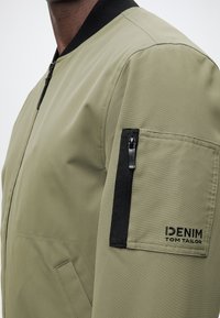 Κοντινή λήψη του μανικιού ενός μπουφάν bomber σε ελαιίον πράσινο με μαύρη τσέπη με φερμουάρ και κέντημα λογότυπου "DENIM TOM TAILOR" στο μπράτσο.