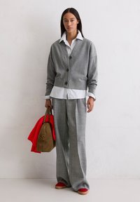 Cardigan gris avec des boutons superposé à une chemise blanche à rayures. Pantalons à carreaux gris, chaussures rouges et un sac marron avec une écharpe rouge.