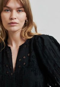 Femme aux cheveux clairs portant de grandes boucles d'oreilles en argent et un chemisier noir texturé à manches bouffantes, le tout sur un fond uni.