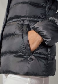 Grå pufferjakke med en glat, skinnende overflade, der har horisontale syninger, sidelomme og en logo-knap i kanten.