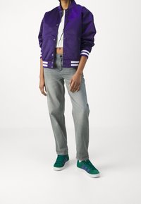 Paarse satijnen bomberjack met witte strepen, grijze vrijetijdbroek en groene sneakers met paarse accenten.