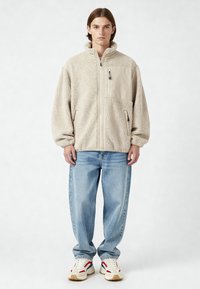 Veste en polaire beige avec col montant, fermeture éclair et poche poitrine, associée à un jean bleu clair et des baskets avec des accents rouges et bleus.