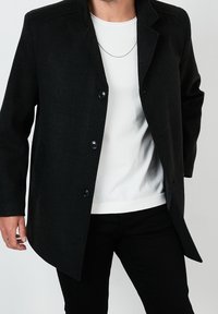 Cappotto nero in lana con un design strutturato, colletto a scendere e chiusure con bottoni. Abbinato a una camicia bianca e pantaloni neri.