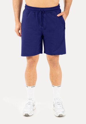 Mand iført marineblå atletikshorts, hvide sokker og hvide løbesko, står med den ene hånd i lommen.