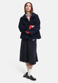Ναυτική ζακέτα από μείγμα μαλλιού με κομψή γραμμή, φαρδιά μανίκια και μπροστινές τσέπες, σε συνδυασμό με φούστα με πιέτες και μαύρες λοφάδες. Ορατές λευκές κάλτσες.