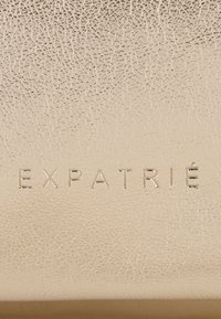 Beżowa skóra o delikatnej fakturze. Wytłoczone litery "EXPATRIÉ" w nowoczesnej czcionce. Gładki, minimalistyczny design, bez dodatkowych wzorów czy akcesoriów.